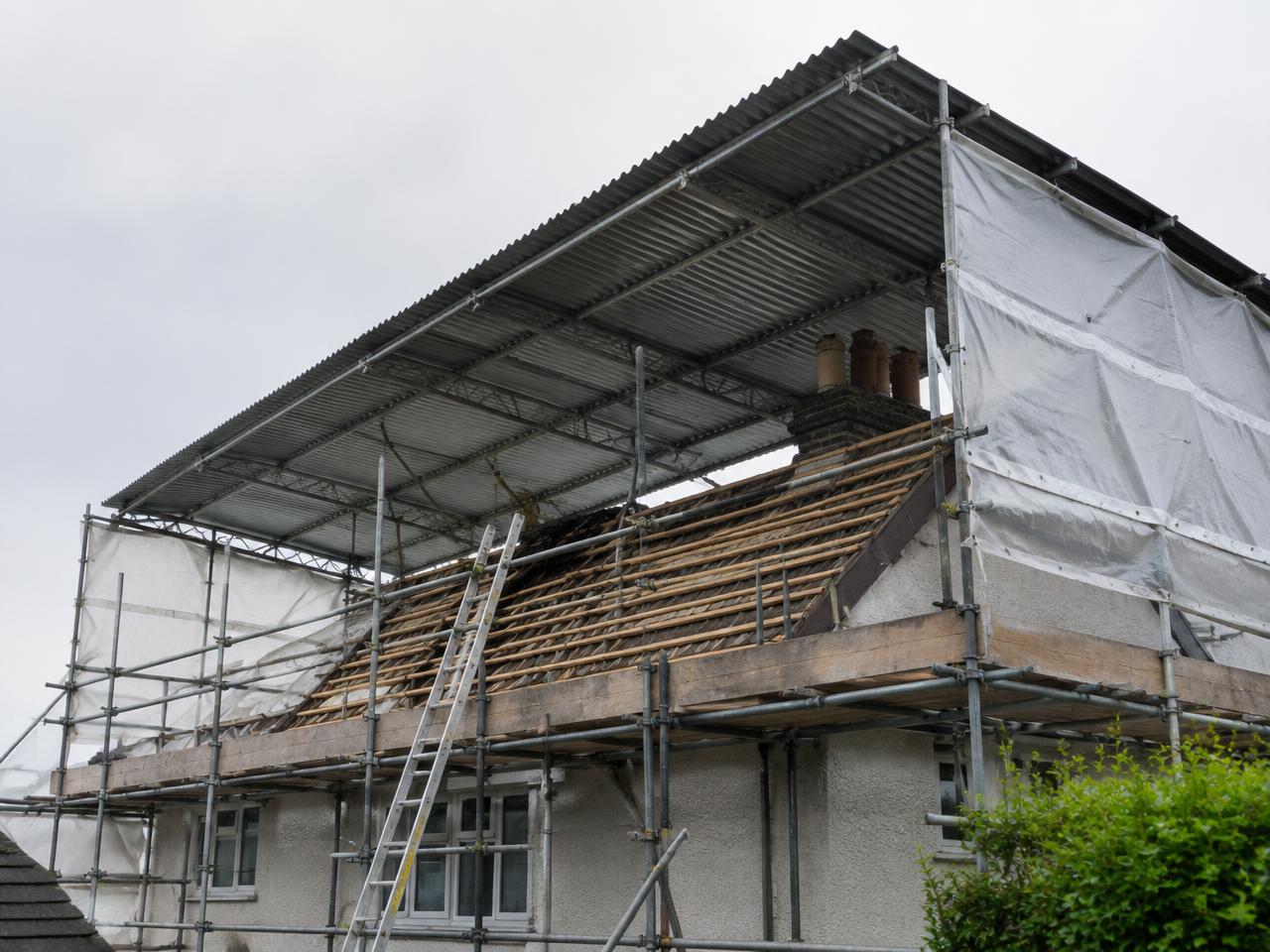 Temporary roof with corrugated sheeting spanning over a stripped house roof, scaffold and shrink wrap to the gable