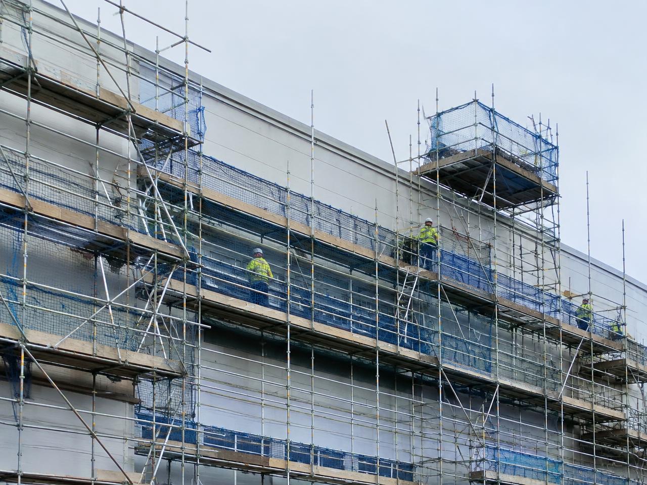 Full-height system scaffold with netting across a large commercial building, workers in hi-vis on the lifts