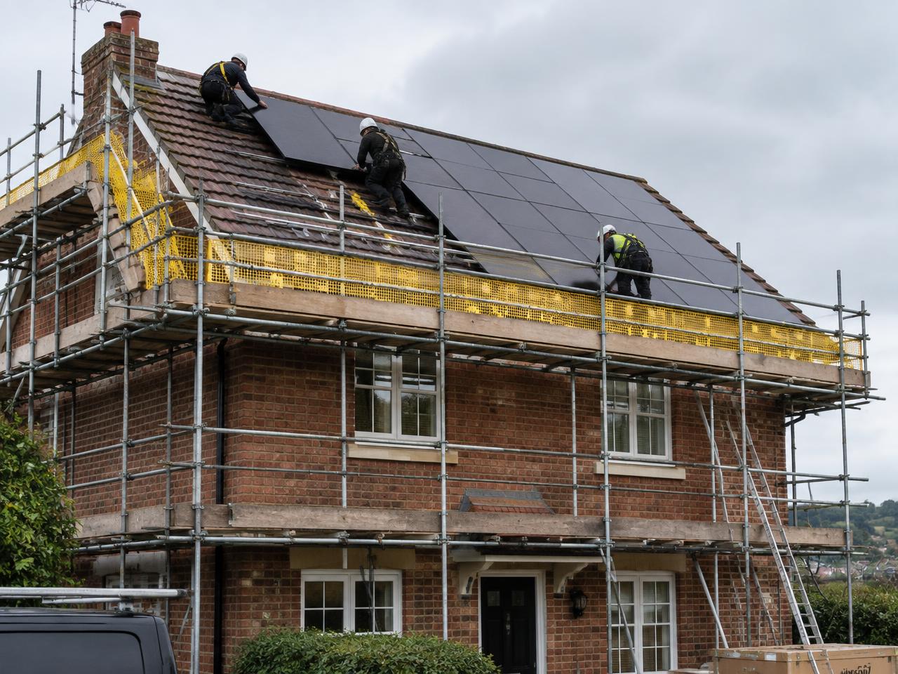 Scaffold with edge protection along the eaves of a brick house while installers fit black solar panels on the roof